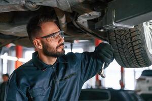 In protective eyewear, under ,vehicle. Car repairman is in the garage with automobile photo