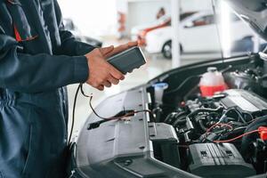 Device for measuring, voltage is in hands. Car repairman is in the garage with automobile photo