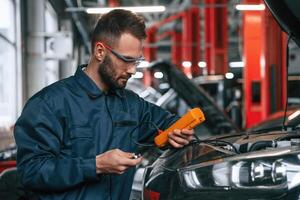Checking electronics by using device. Car repairman is in the garage with automobile photo