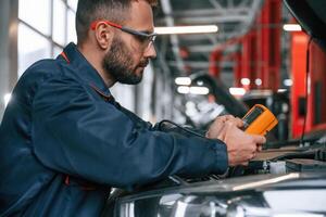 Yellow colored device for measuring ,electricity is in hands. Car repairman is in the garage with automobile photo