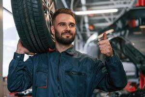 Holding a ,,,tire. Car repairman is in the garage with automobile photo
