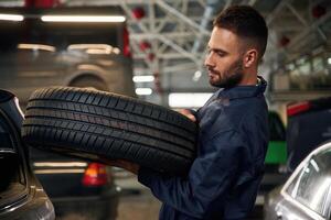 Holding ,a ,tire. .Car repairman is in the garage with automobile photo