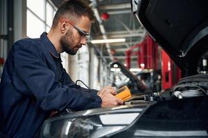 Yellow ,colored device for measuring electricity is in hands. Car repairman is in the garage with automobile photo