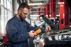 Professional electro, device in hands. Car repairman is in the garage with automobile photo