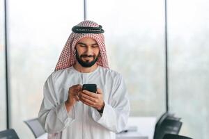 With smartphone in hand.. Successful Muslim businessman in traditional outfit in his office photo