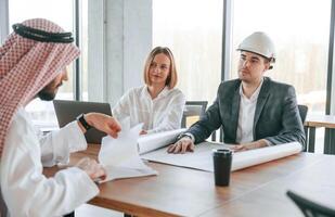 With architect sitting by,, the table. Muslim businessman in traditional outfit with colleagues in office photo