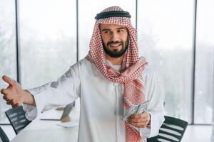 Holding money ,in hands. Successful ,Muslim businessman in traditional outfit in his office photo