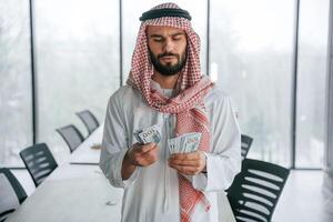 Holding money in, hands. Successful Muslim businessman in traditional outfit in his office photo