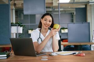 Nutritionist, with apple and laptop. Female doctor in white coat is indoors photo