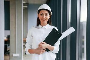 Plan and, notepad ,,in hands. Female architect in hard hat and in white clothes is indoors photo