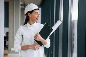 Plan and notepad in hands... Female architect in hard hat and in white clothes is indoors photo