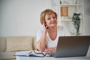 Front view. Sitting ,by table with laptop. Senior elderly woman is at home in the living room photo
