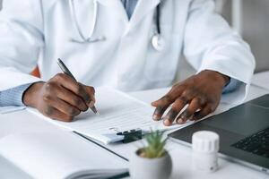 Filling in the document, writing by using pen. Black man as doctor in the clinic office photo