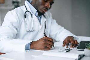 Writing information into the document.. Black man as doctor in the clinic office photo