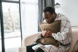 Sitting on sofa,, holding pillow. Sick black man is at home photo