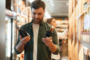 Guy, holding two bottles in hands. Man and woman are choosing wine in the shop photo