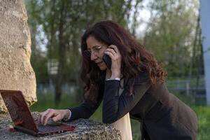 beautiful middle-aged businesswoman talking on cell phone and working on laptop leaning on concrete cube as desk outdoors photo