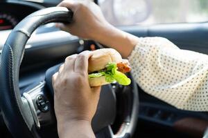 Asian woman driver hold and eat hamburger in car, dangerous photo