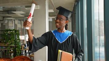African American graduate in cap and gown proudly holds diploma and books while standing in a bright modern academic space video