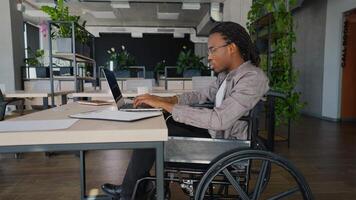 African American freelancer programmer in a wheelchair coding on a laptop at a desk in a modern coworking space video