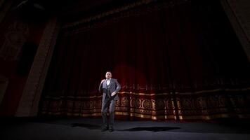 masculino anfitrión hablando en escenario. elegante hombre en gris disfraz ejecutando en grande escena usos gestos actor Moviente alegremente en escenario. video