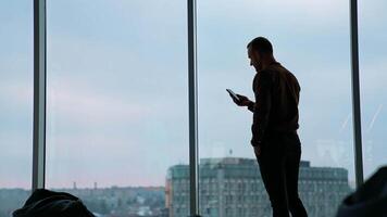 Office worker with a phone. Young businessman looking into the phone near the window in office on city buildings background. Panoramic window view. video