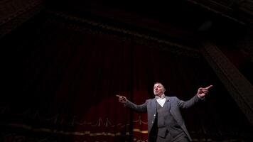 Actor performing on stage. Man in grey suit speaking with many gestures on red theatrical curtains background. View from below. video