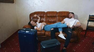 Man and woman rest on a couch tired from journey. Young couple lying on sofa waiting for the leaving the hotel on suitcases packed background. video