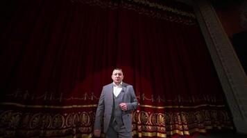 Man in gray costume on stage. Middle-aged actor talking on camera on luxury red curtains background in the theater. Slow motion. video