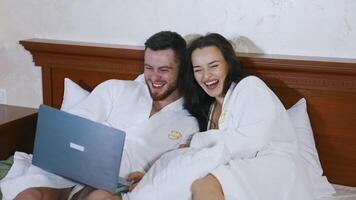 Joyful young couple on bed. Handsome man and attractive woman in white bathrobes laying on bed and laughing while looking into laptop. Happy together. video
