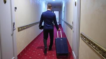Man in suit with suitcase in hotel. Back view of a businessman with a trolley bag walking to his room in the hotel corridor. video