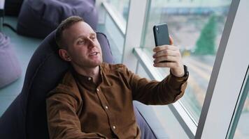 Man is making a call. Handsome young businessman sitting in a soft chair near the window and talking through the phone in office. video