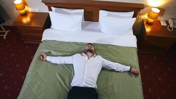 Tired businessman on comfortable bed. Man in white shirt and trousers laying on bed relaxing in hotel after the hard journey. Top view. video