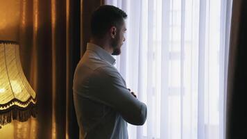 Handsome man near the window. Young businessman in white shirt looking on the window view standing near the large lamp in the hotel. Business trip. Side view. video