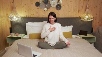 Comfortable woman in pajamas follows an online meditation class in her bedroom, sitting in lotus position on her bed with a laptop in front of her video