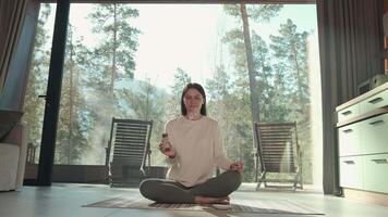 Young woman practicing meditation with incense in a modern home with large windows overlooking a peaceful forest, creating a serene and tranquil atmosphere video