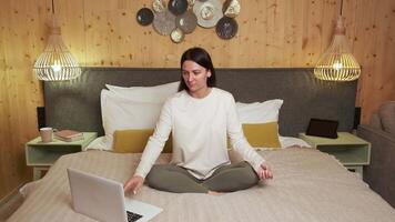 Serene woman meditating in lotus position on bed, using laptop in soft-lit wooden bedroom, embodying mindful wellness and digital learning during peaceful home practice video