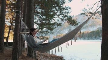 Young woman freelancer is working remotely lying in hammock between trees on snowy shore of frozen lake, enjoying beautiful winter landscape video