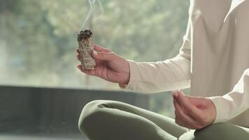 Wellness practitioner meditates in lotus position while holding a smoldering white sage bundle, using it for spiritual cleansing and energy purification video