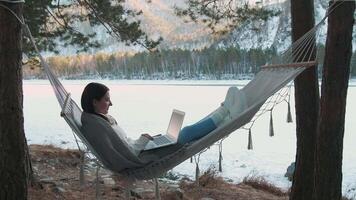 Freelancer woman working remotely on laptop while relaxing in hammock between trees, overlooking frozen lake amid snowy winter forest landscape video