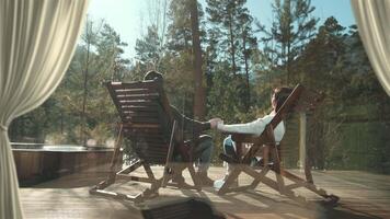 Young couple enjoying a relaxing moment, holding hands while sitting in rocking chairs on a wooden deck, admiring a mountain view from a resort video