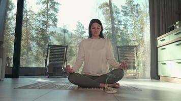Peaceful woman practicing yoga and meditation in modern home interior, finding inner tranquility while overlooking serene forest landscape through large window video