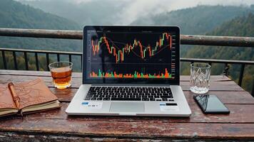 Laptop computer displaying stock market chart, whiskey glass, and smartphone on wooden table with notebook, set against a mountain view, remote work, online trading and financial video
