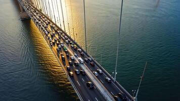 Aerial view of suspension bridge filled with vehicles during golden sunset, reflecting on water surface, showing urban traffic, transportation, architecture, and daily commute in modern video