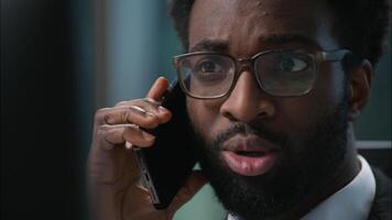 Close up face professional worker African American man businessman talking mobile phone cellphone call consulting client male business manager employee guy looking at monitor computer solve in office video