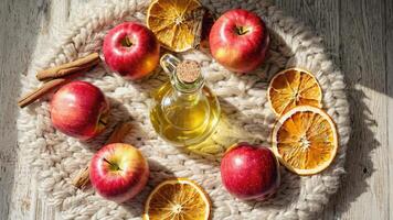 Top view of a glass bottle filled with golden apple oil, surrounded by red apples, dried orange slices, and cinnamon sticks on a cozy knitted blanket, creating a warm, rustic, and video