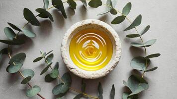 Bowl of golden essential oil placed at the center of a eucalyptus leaf circle on a light stone surface, symbolizing natural wellness, aromatherapy, purity, health care and holistic self video