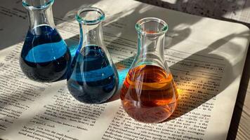 Three flasks filled with blue and orange liquid are placed on open old retro book under sunlight, casting colorful reflections and shadows on the pages of scientific academic alchemy video