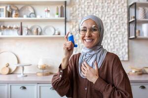 Portrait of smiling young Muslim woman in hijab holding asthma and allergy inhaler at home in kitchen, pointing and posing to camera. photo