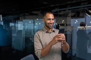 Smiling african american businessman inside office in shirt using phone, man typing message and browsing internet pages online standing near window at workplace in the evening. photo